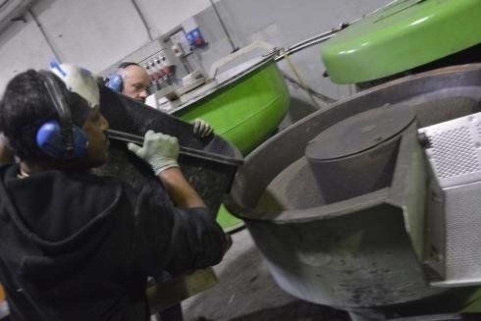 Due operai in una fabbrica caricano un grande contenitore sui macchinari, indossando protezioni acustiche.