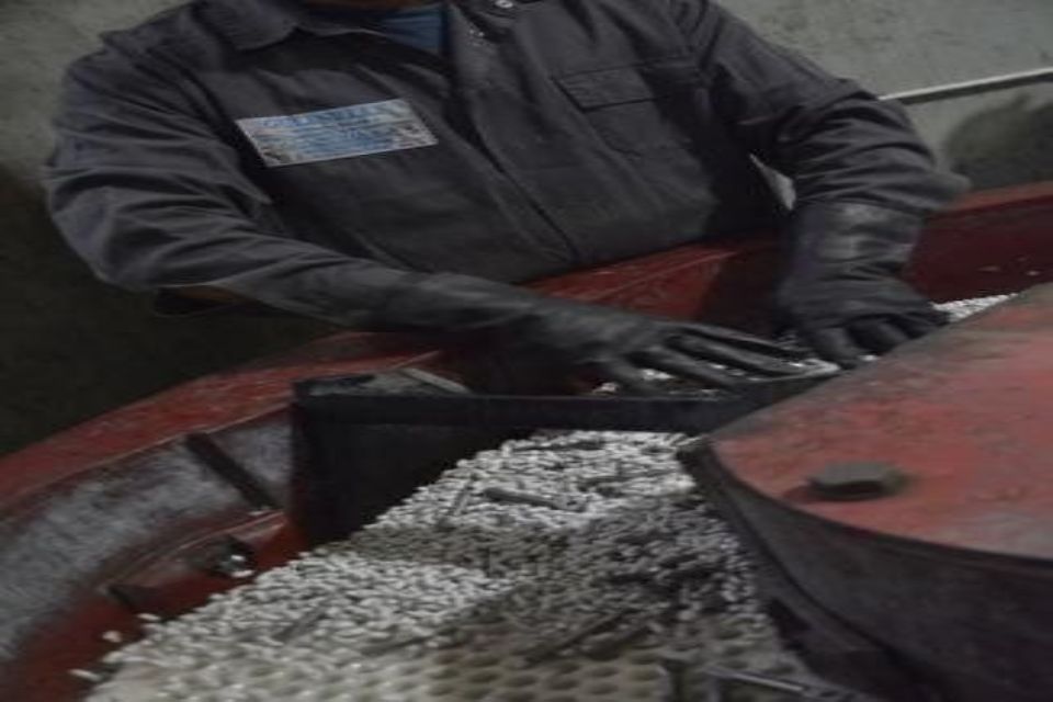 Persona in abiti da lavoro e guanti che ispeziona i componenti in un contenitore contenente piccoli granuli abrasivi bianchi.