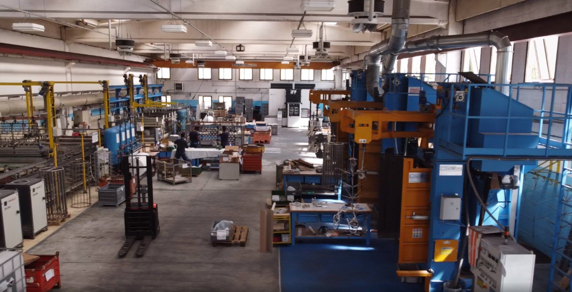 Reparto di fabbrica con grandi macchinari blu e un carrello elevatore che sposta i materiali.