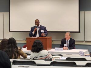 Man speaking at podium, another at table. Audience in foreground, screen behind.