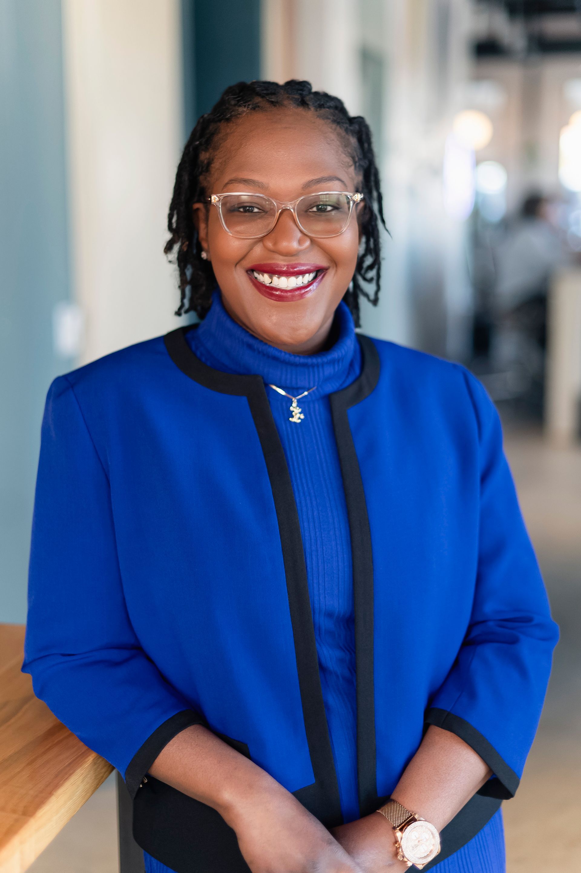 Woman in blue jacket, turtleneck, glasses smiles, leaning on wooden surface.