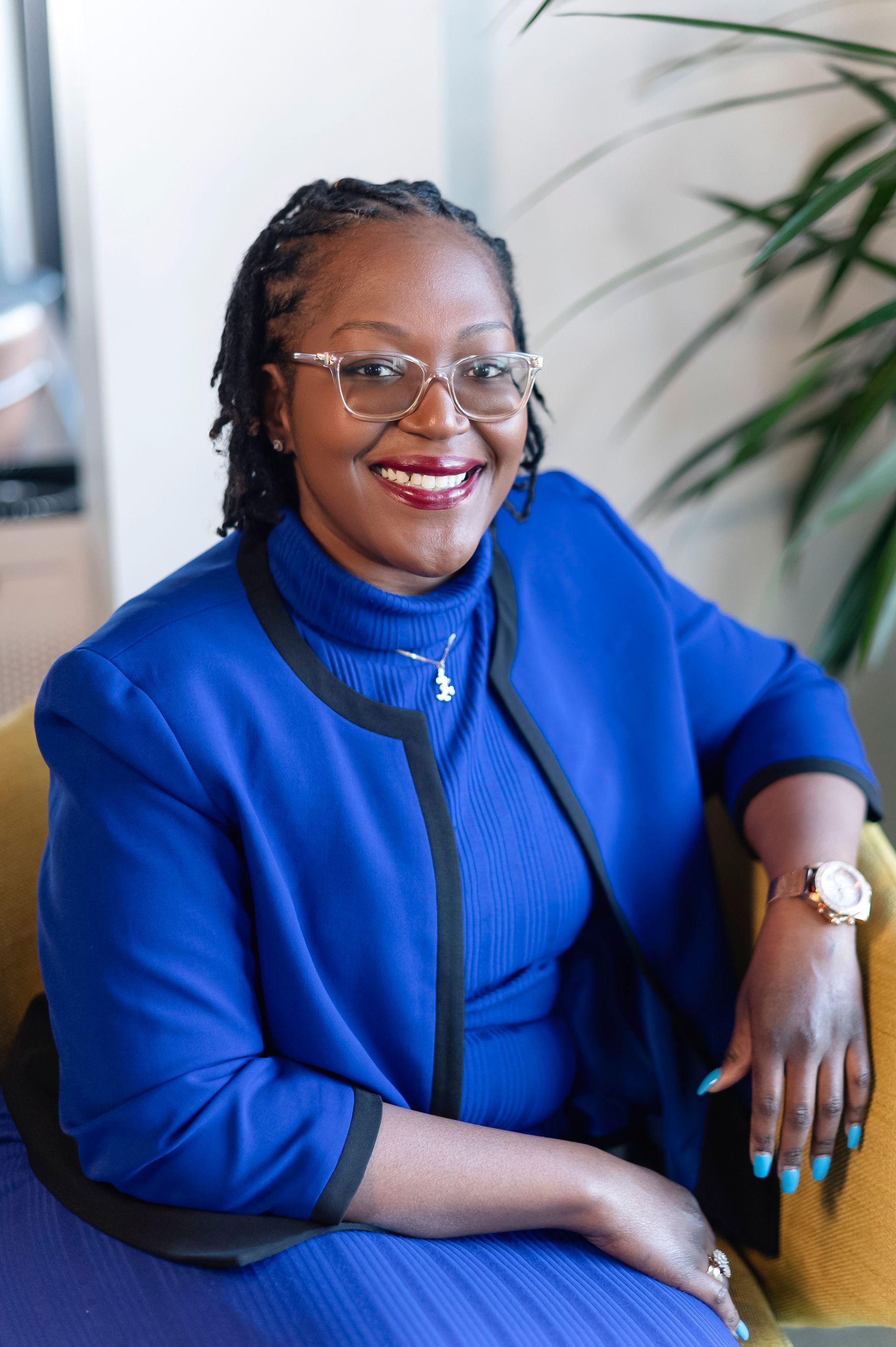 Woman with glasses smiles while sitting in a yellow chair, wearing a blue blazer.