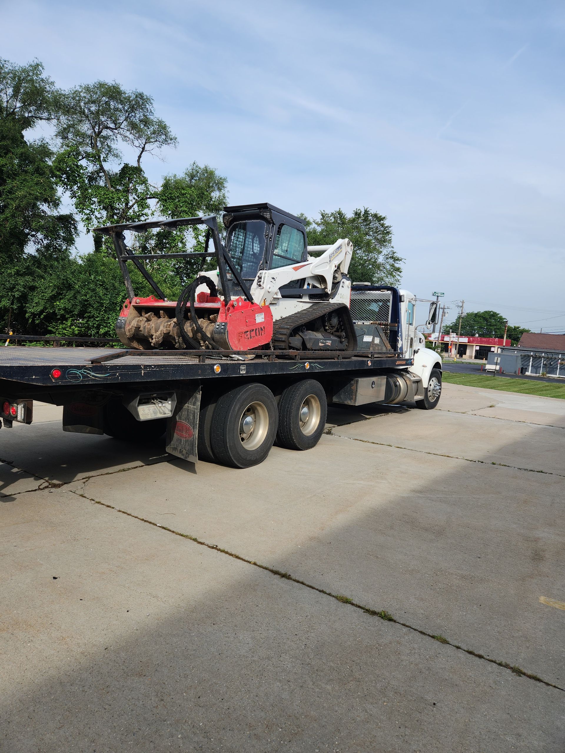A white tracked skid steer loader with a red forestry mulcher attachment parked on a flatbed tow truck on a paved lot.