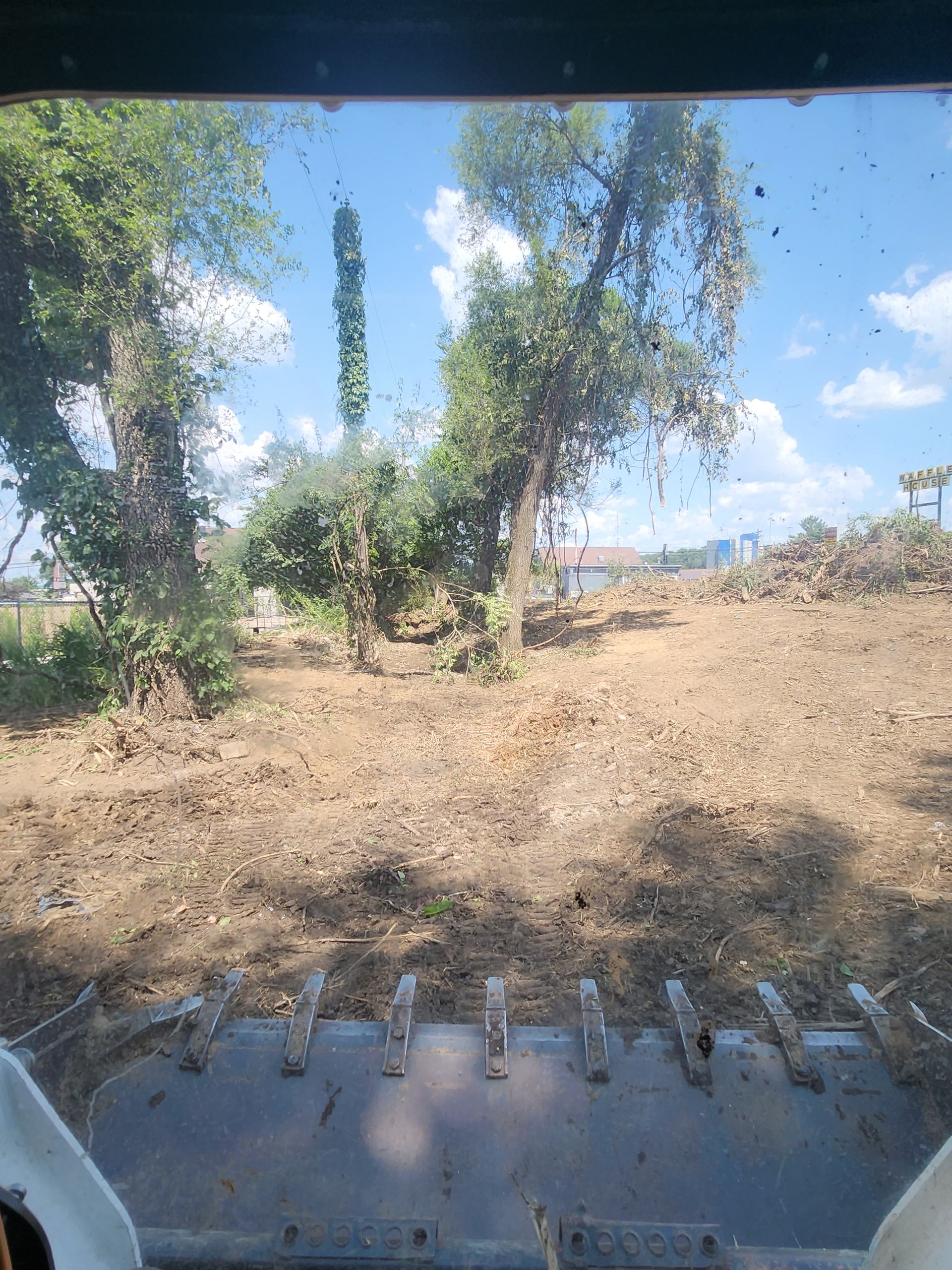 View from the cab of a skid steer or loader showing a cleared, dirt-covered lot with trees under a blue sky.