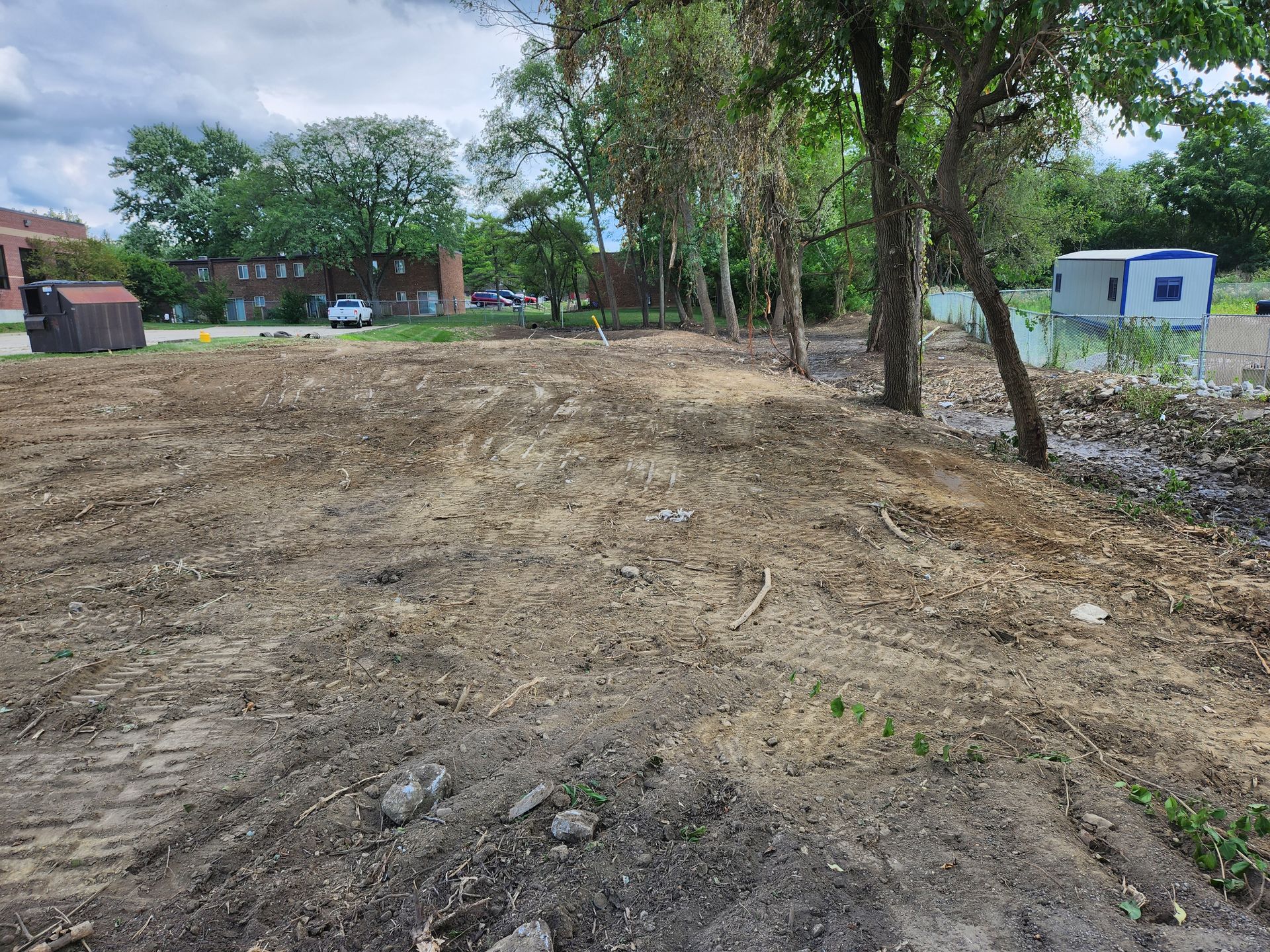 A cleared, dirt-covered construction site with a row of trees, a small shed, and distant buildings under a cloudy sky.