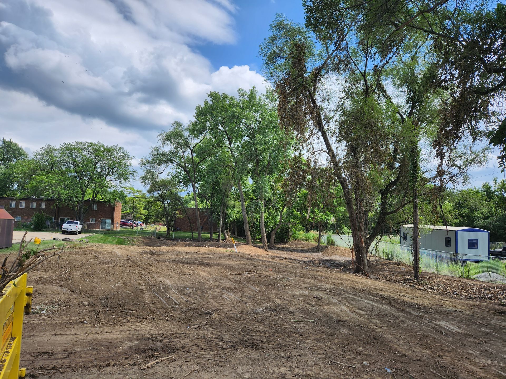 Cleared dirt lot with trees and buildings in the background, under a bright, cloudy blue sky.