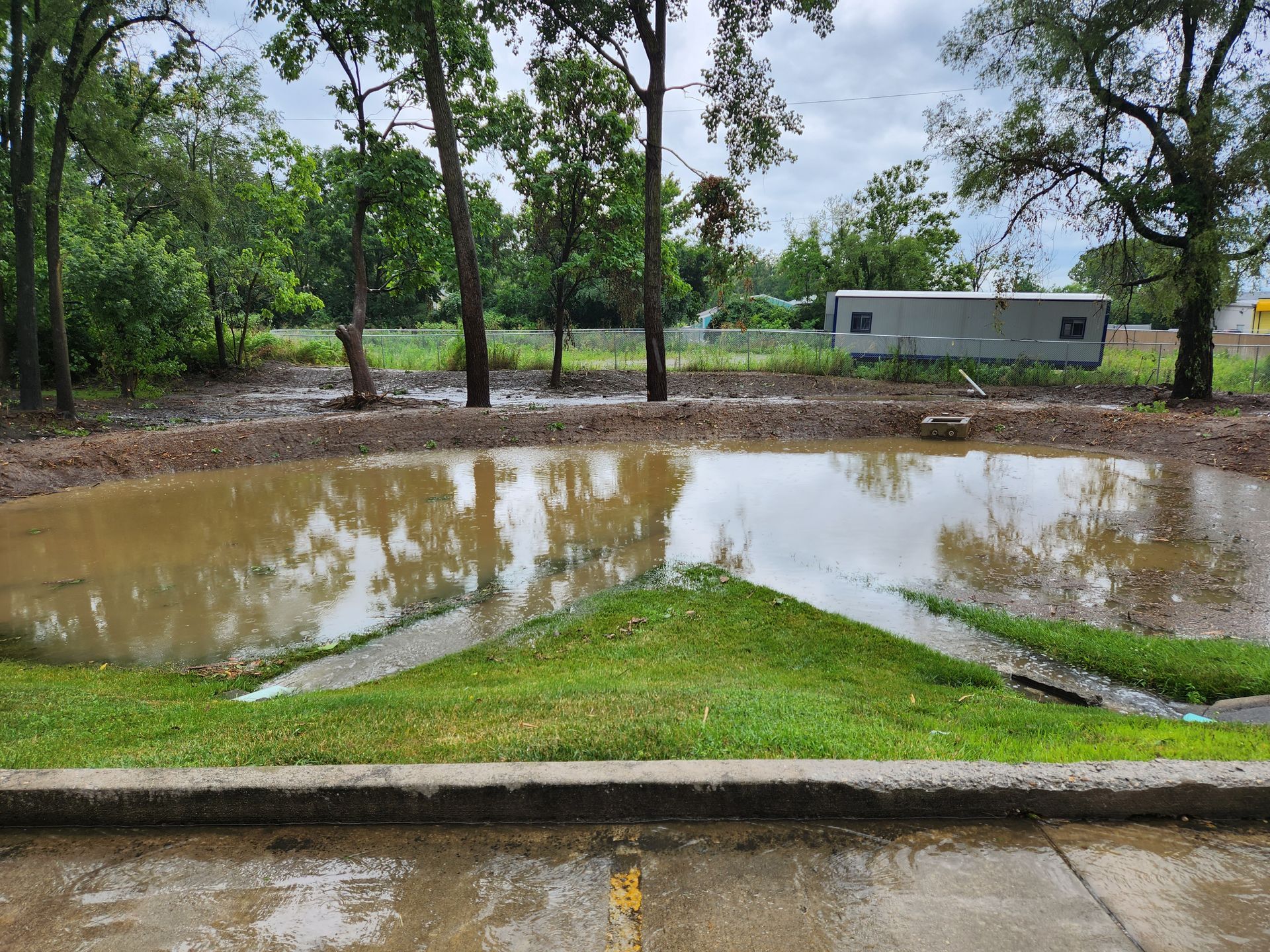 A muddy pond reflecting trees, adjacent to a grassy embankment and a paved area under a cloudy sky.