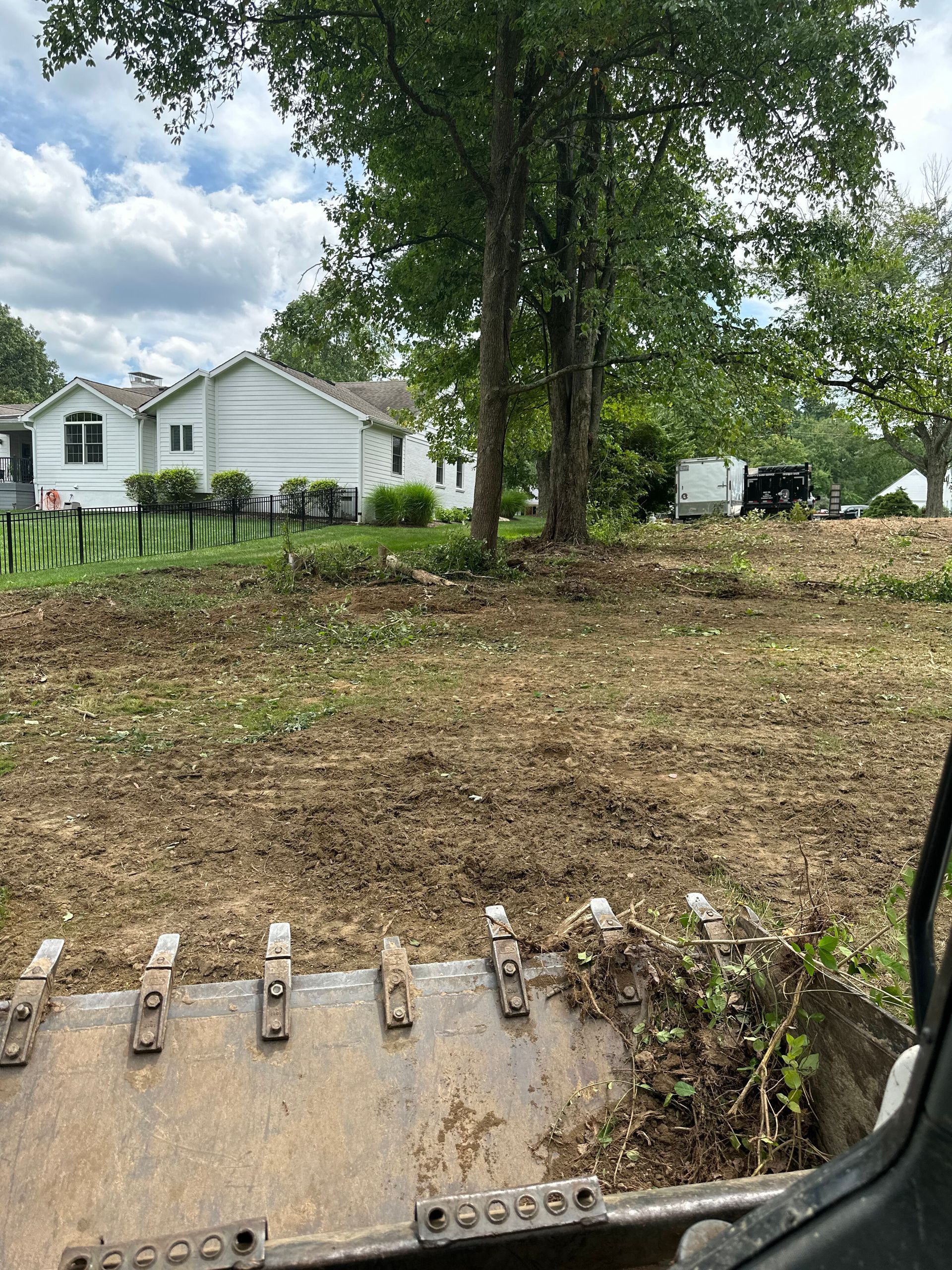 View from a skid steer loader bucket looking at a freshly cleared dirt lot with trees and a white house in the distance.