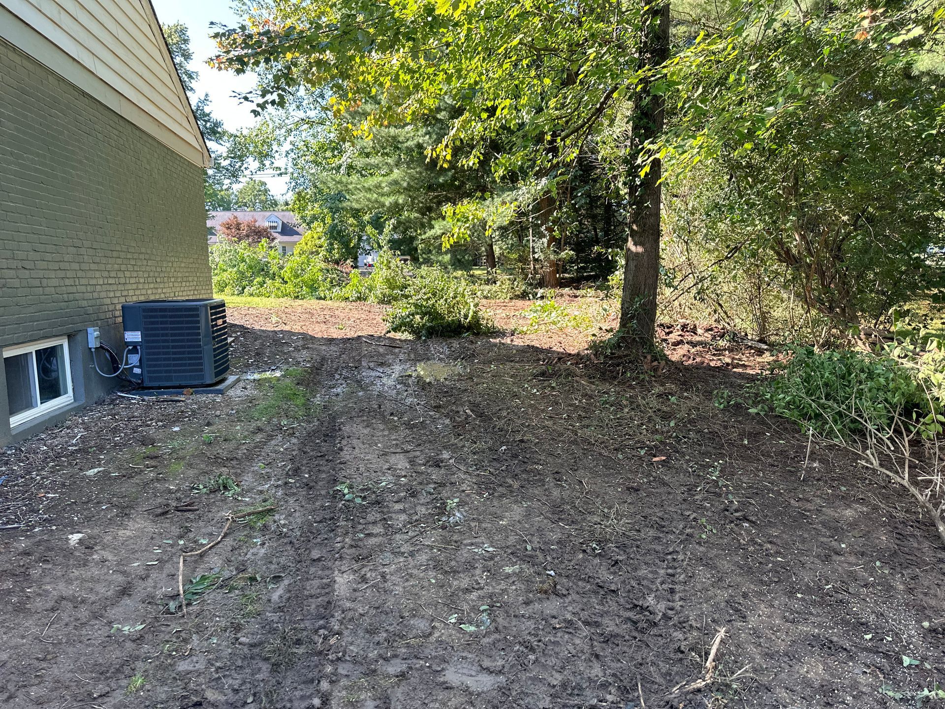 A side yard with cleared, bare soil next to a beige brick house and a dark grey HVAC unit, framed by mature trees.