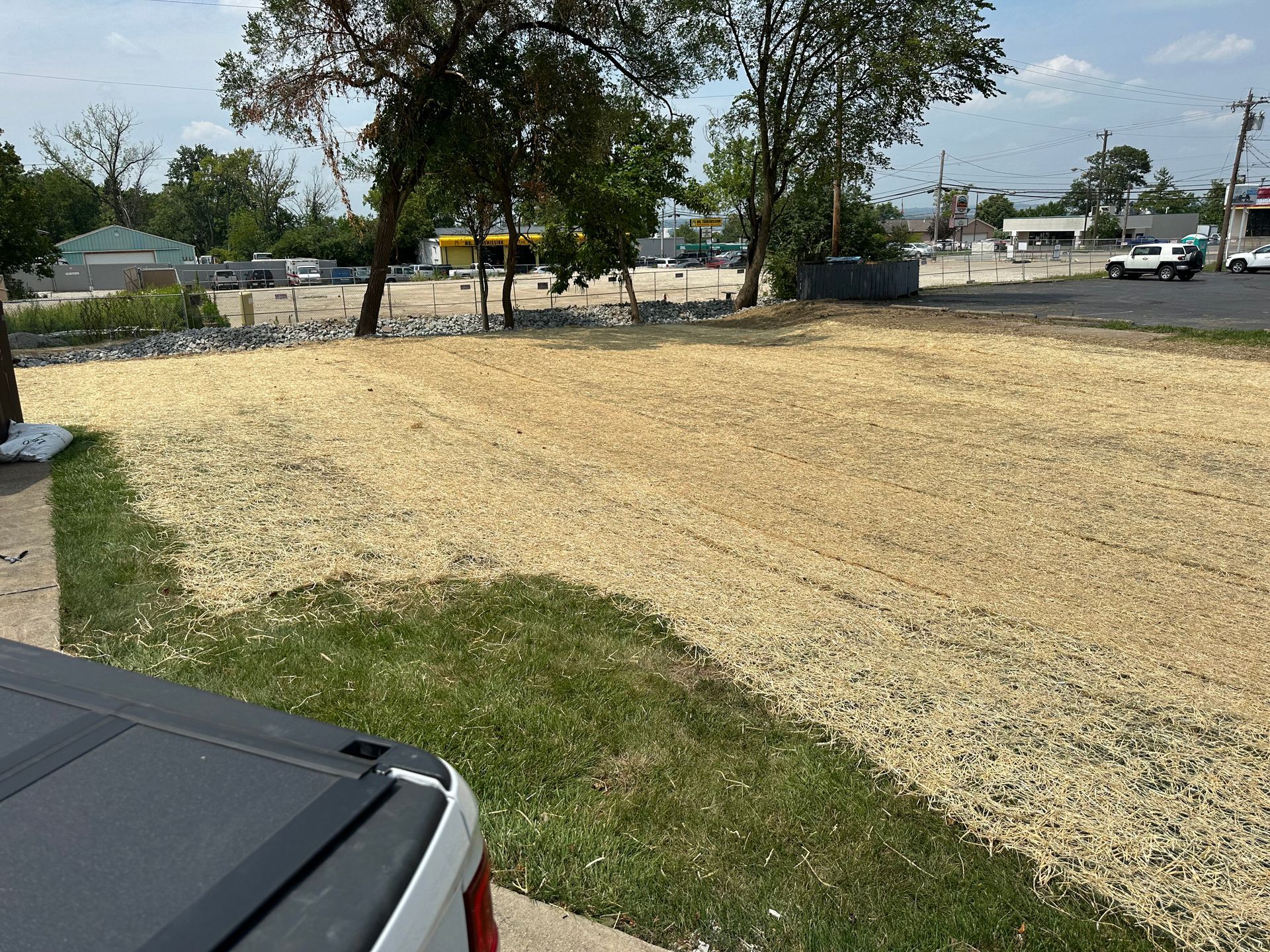 A view from a vehicle of a cleared, gravel-covered lot bordered by green grass and trees under a sunny sky.