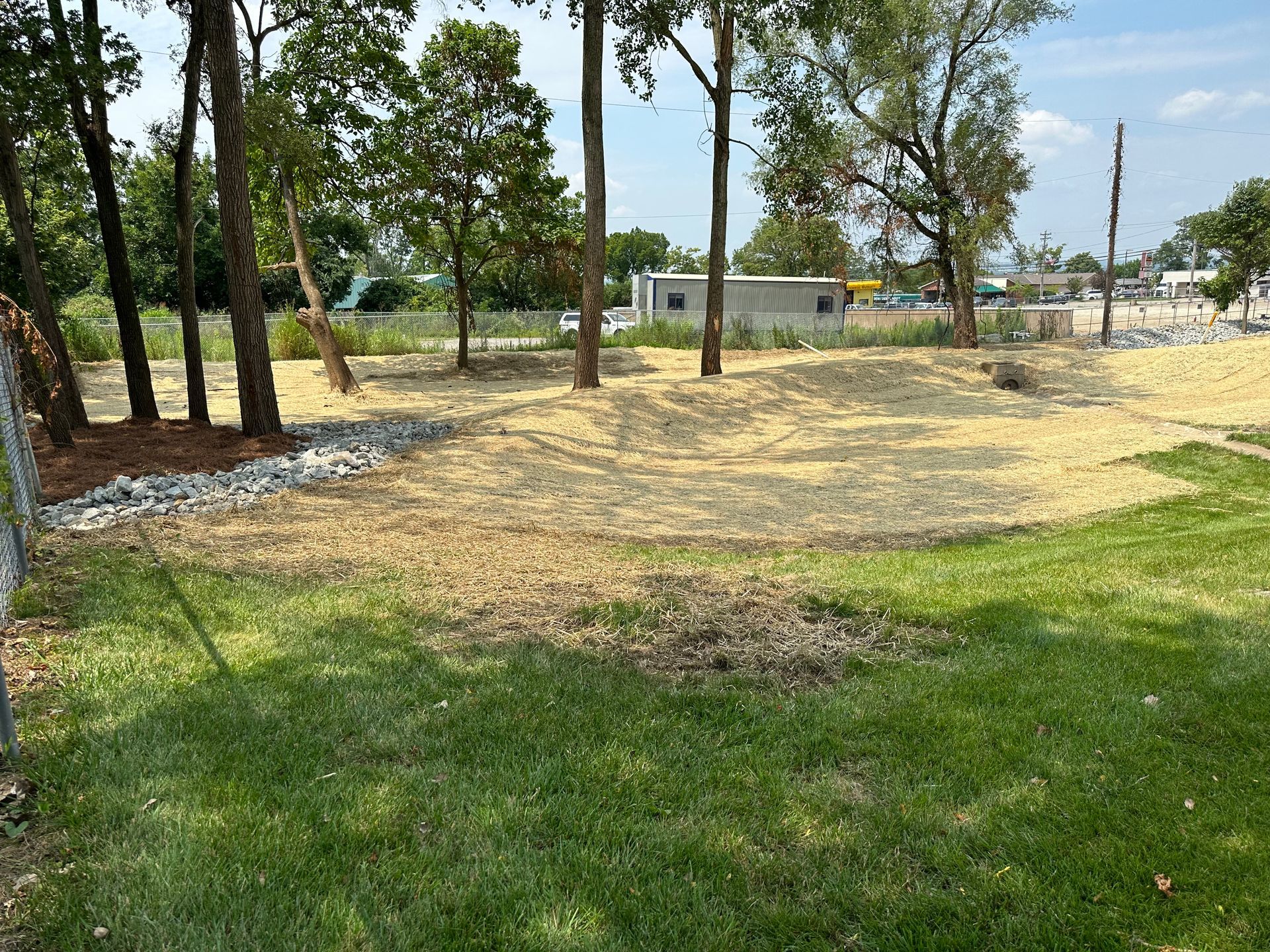 A grassy foreground leads to a dirt embankment with small trees and rocks, backed by a building under a bright blue sky.