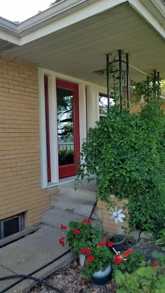Red front door with steps, surrounded by tan brick and foliage.