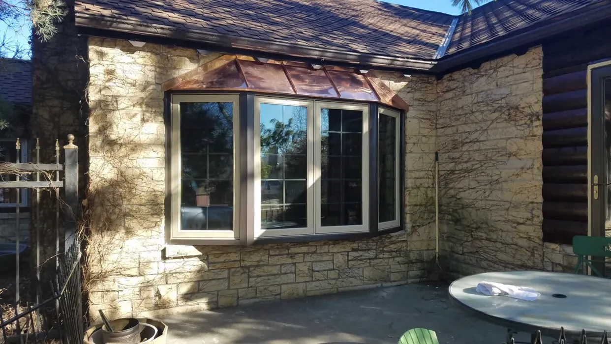 Bay window with copper roof on a stone house, next to a patio with a table.