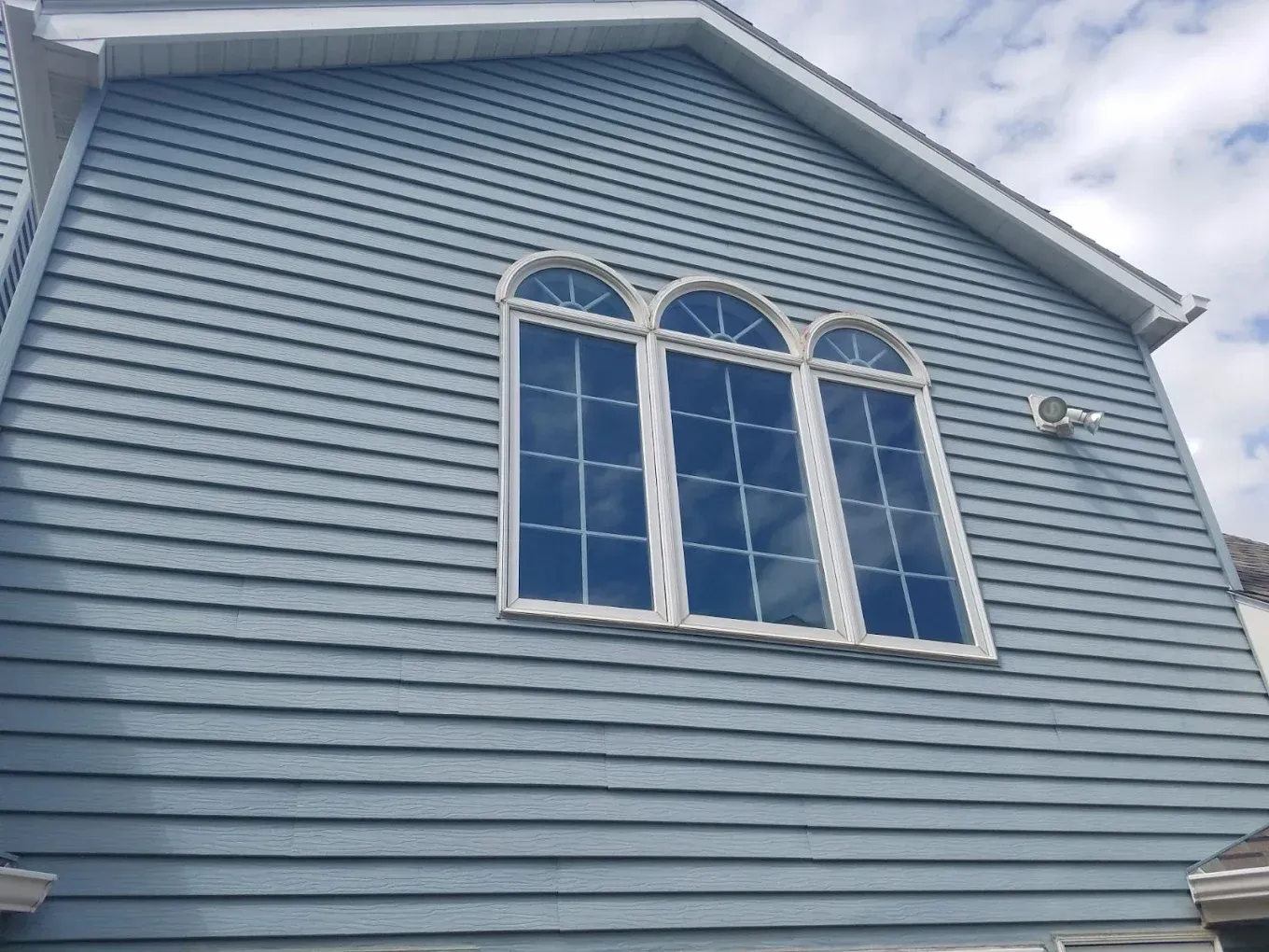 Blue siding on a house with white trim and three arched windows reflecting a cloudy sky.