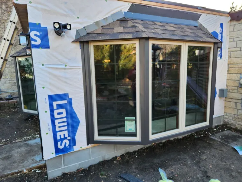 Bay window under construction, blue Lowe's wrap. Gray trim, brown roof. Exterior with floodlights and stone foundation.