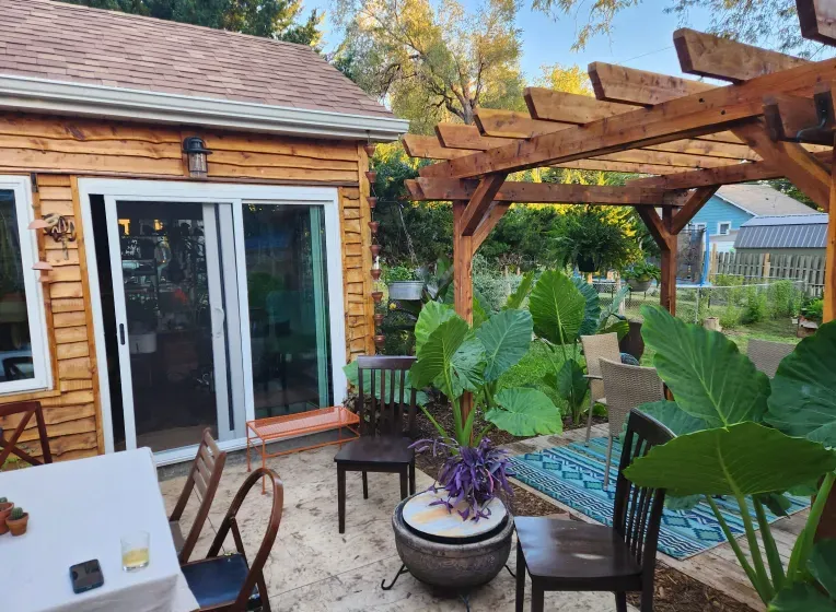 Outdoor patio with wooden pergola, seating, and large-leafed plants. Building has sliding glass door.