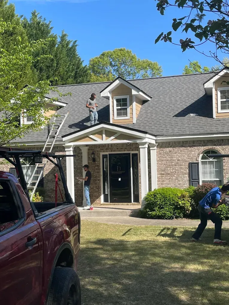 Workers repairing a roof on a brick house. One man is on the roof, two are on the ground. Sunny day.