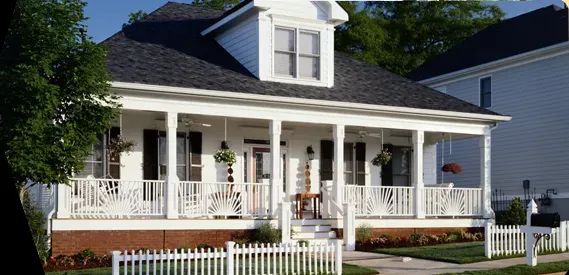 White house with black roof, large porch, white picket fence, and greenery.