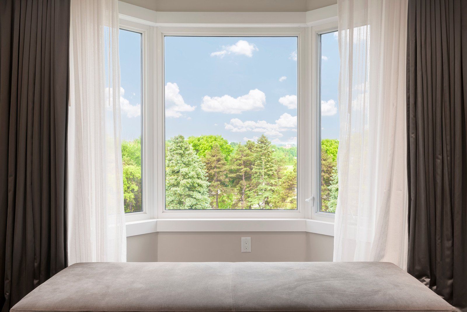 Bay window with a view of trees and a blue sky, framed by white and dark curtains, with a bench in front.