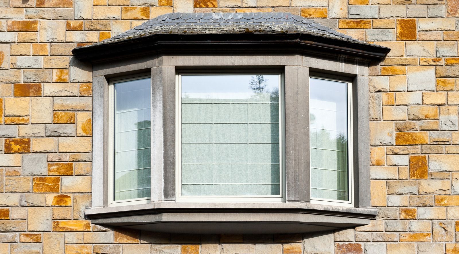 Bay window with gray stone trim on a brick building; white blinds cover the glass.