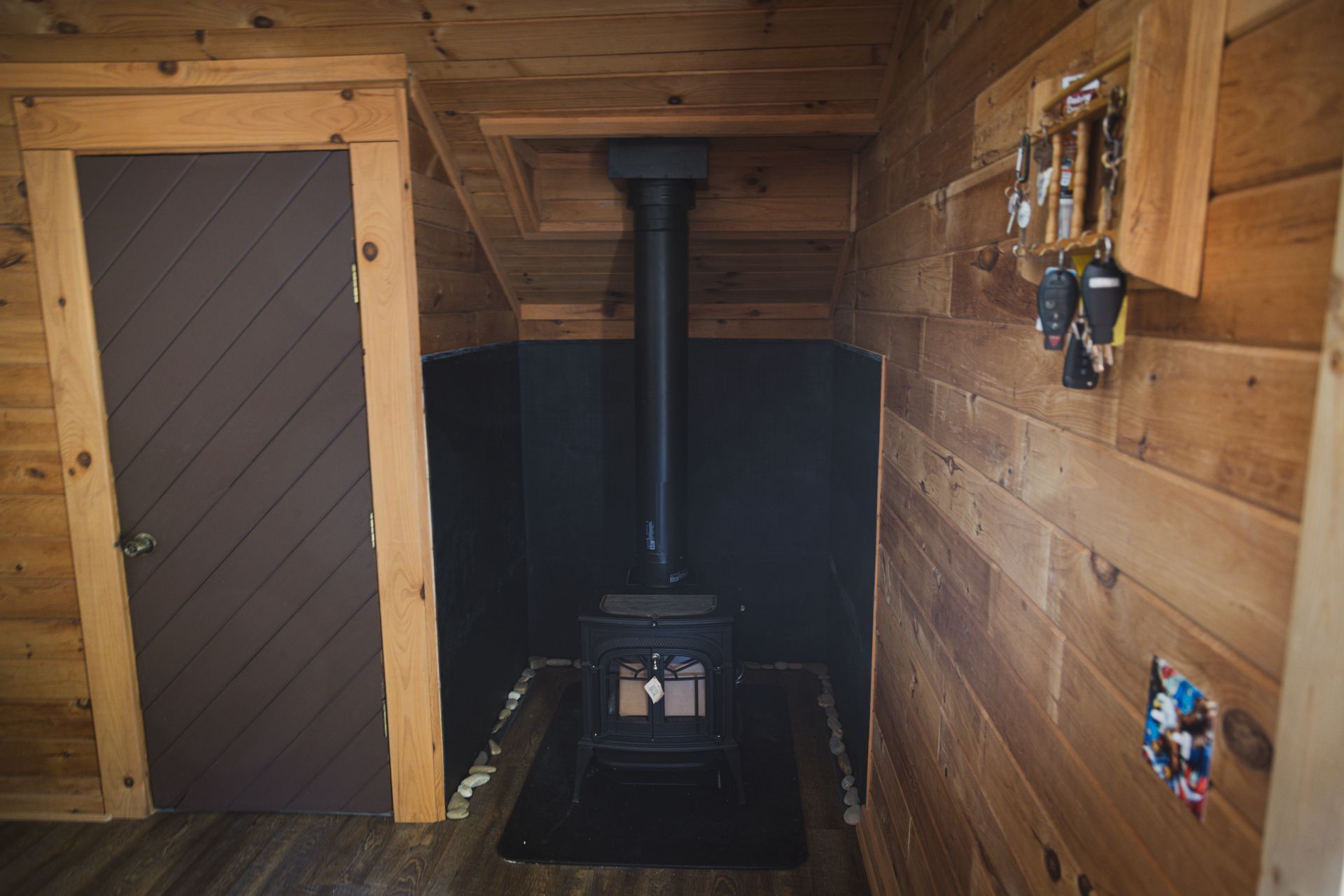 Cozy cabin interior with a black wood-burning stove and a brown door. Wooden walls, a key rack, and a dark-painted alcove.