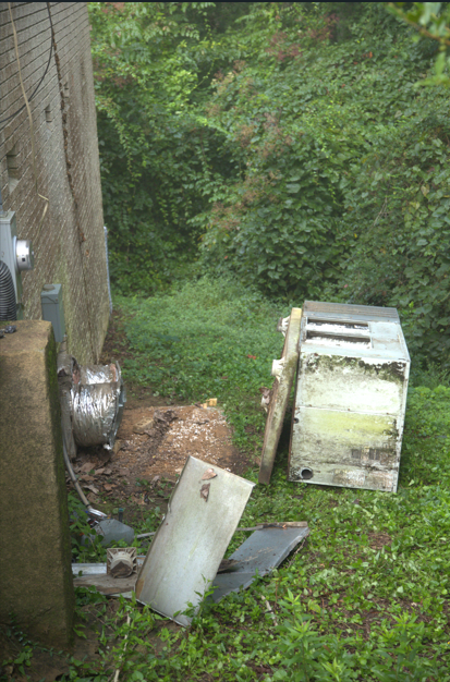 Old, damaged appliance parts and metal debris on overgrown grass next to a building and foliage.