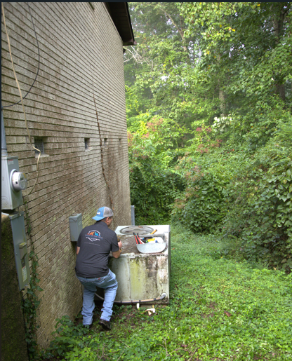 A person works on an old, white appliance next to a building; overgrown foliage is in the background.