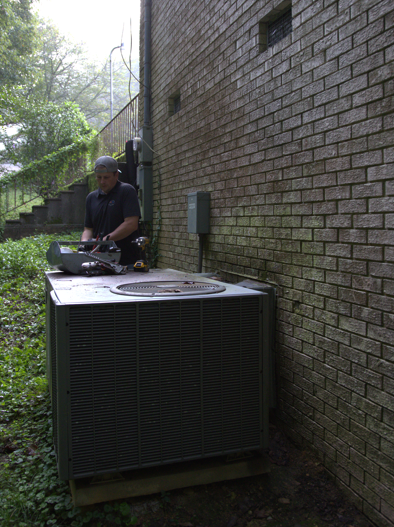 HVAC technician working on an air conditioning unit near a brick building.