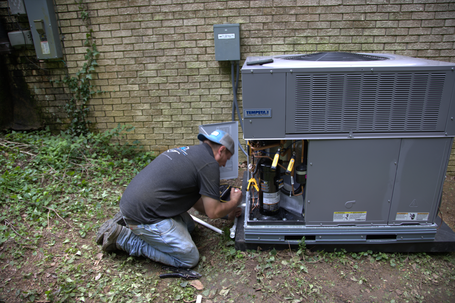 HVAC technician kneels, working on an air conditioning unit outside near a brick wall.