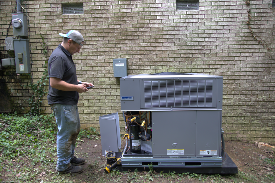 Man working on an outdoor HVAC unit, checking it with a tool in hand near a brick building.