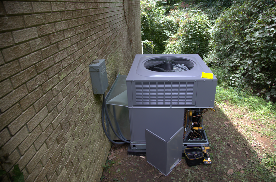 Air conditioning unit near a brick wall and foliage.