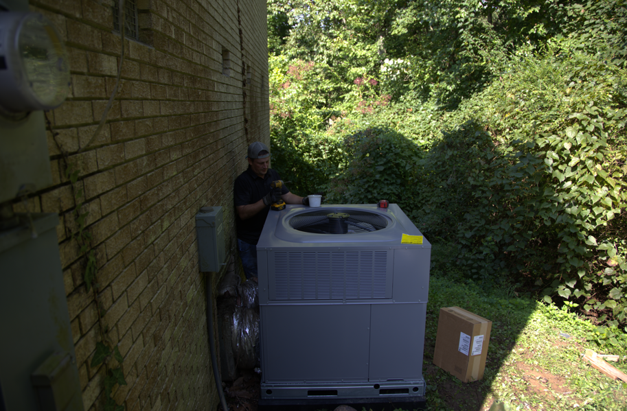 Man working on an air conditioning unit outside a brick building, surrounded by greenery.