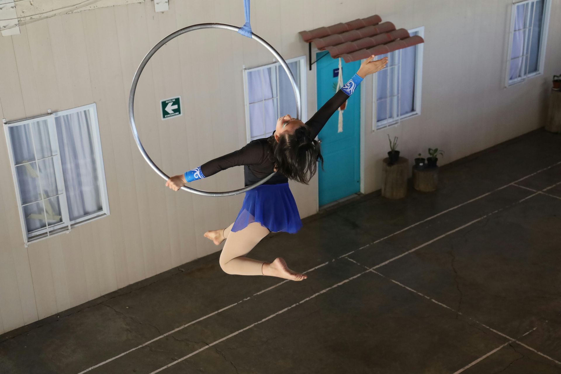 A performer in a black top and blue skirt hangs in an aerial hoop, posed with arms extended in a bright indoor room.