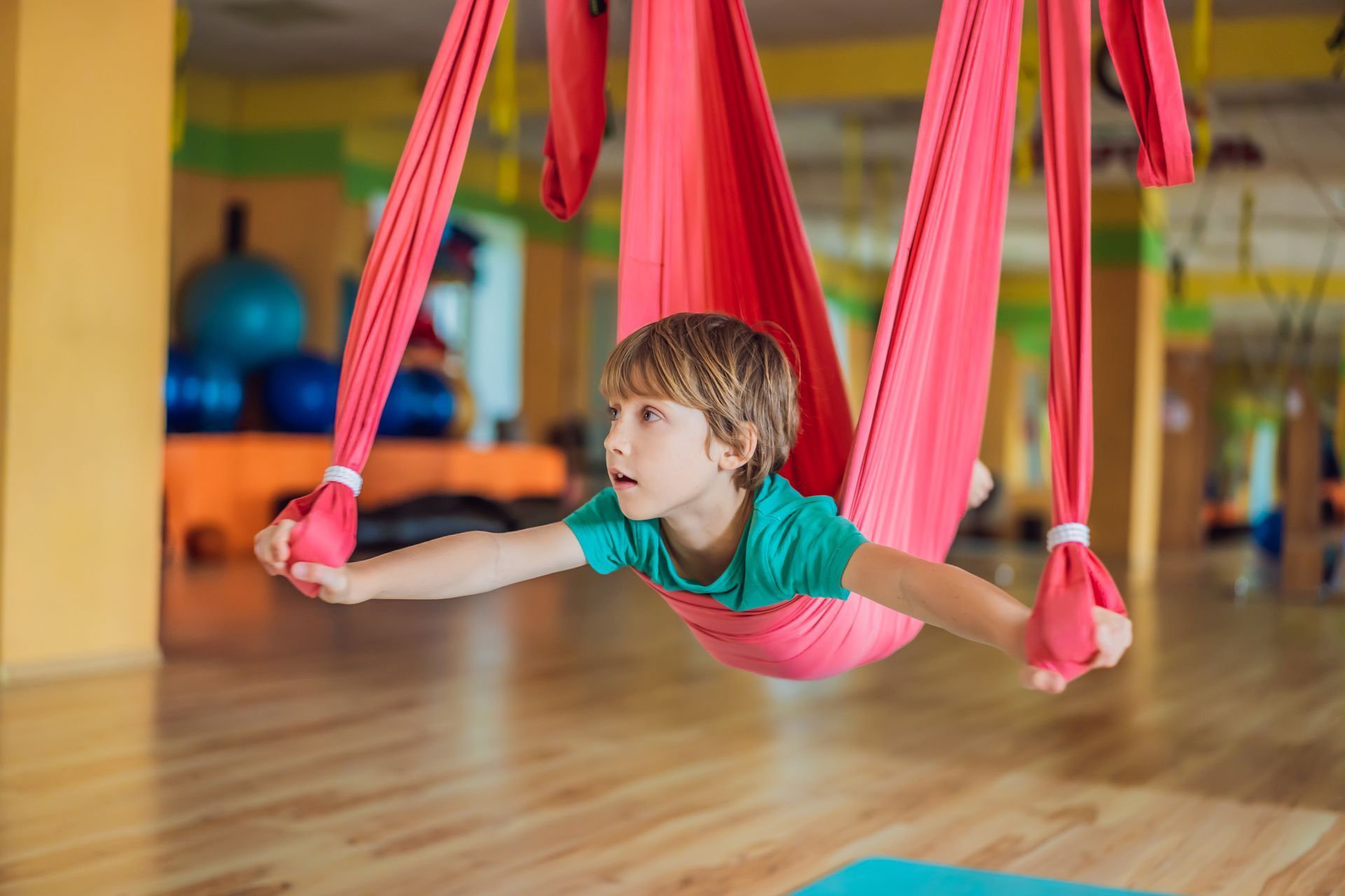 A child in a green t-shirt performs aerial yoga, suspended in a bright red fabric swing inside a gym.