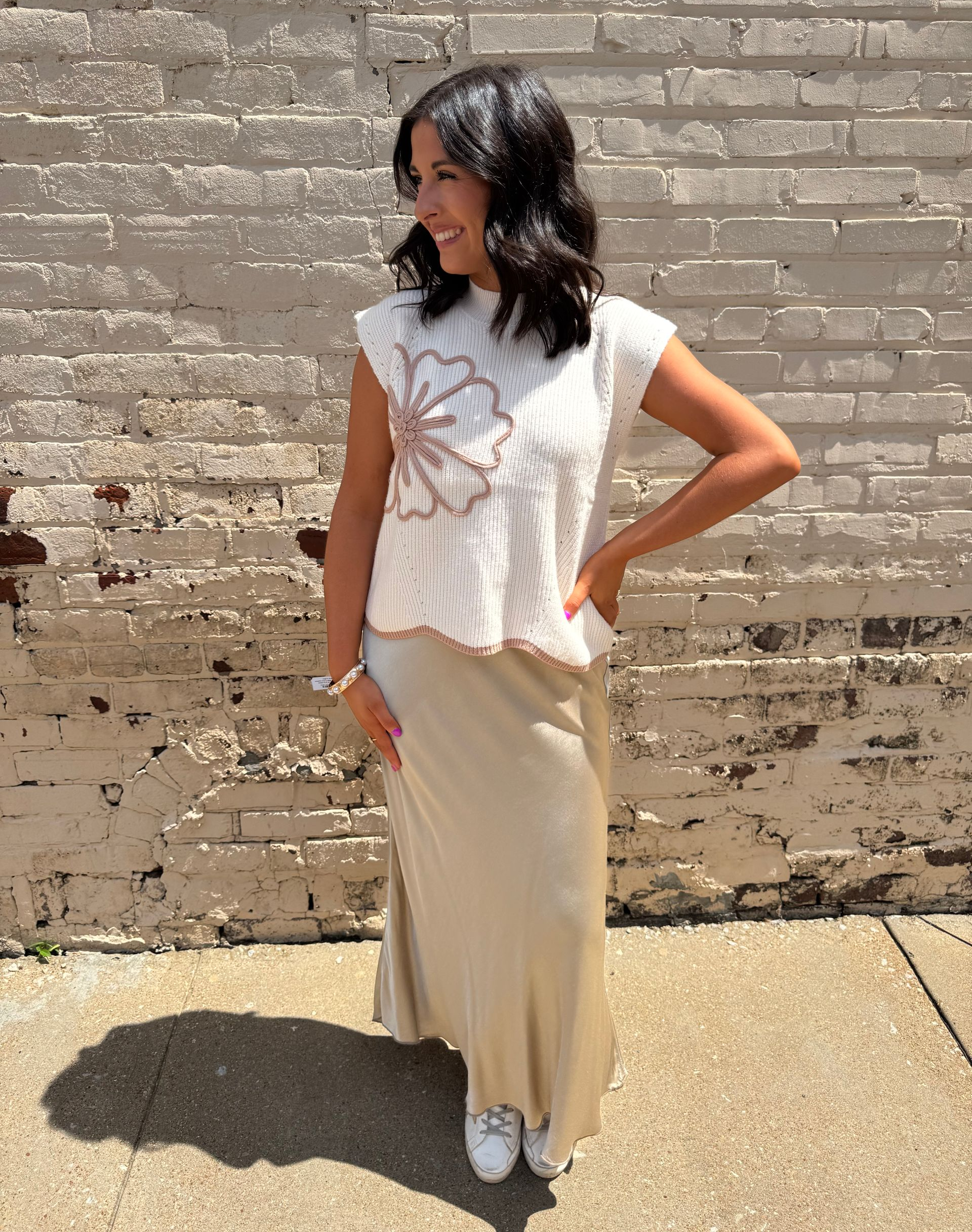 Woman in cream top with flower design and long gold skirt smiles near a brick wall.