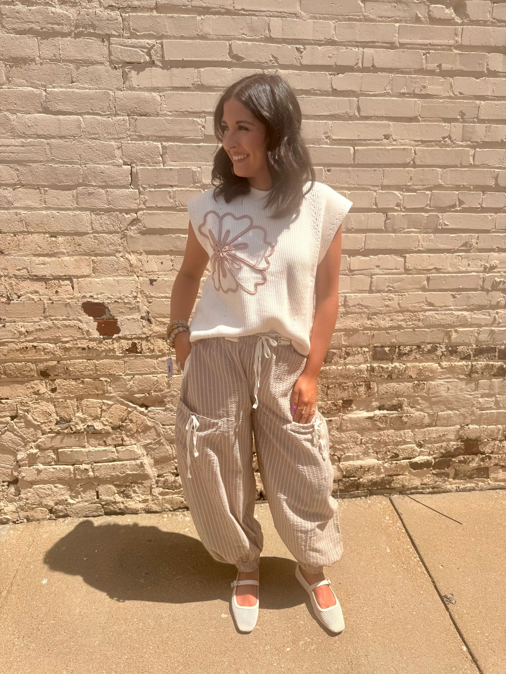 Woman in white floral top and patterned pants stands in front of a brick wall.