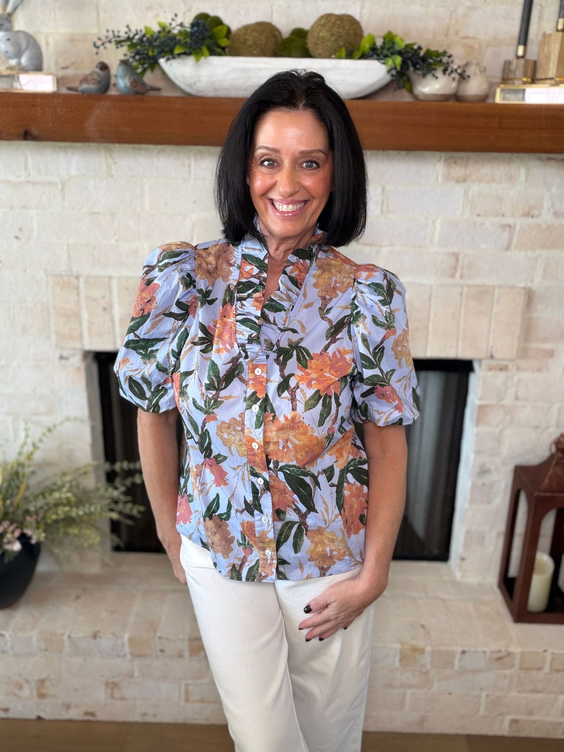 Woman in blue floral top and white pants stands in front of a brick wall.