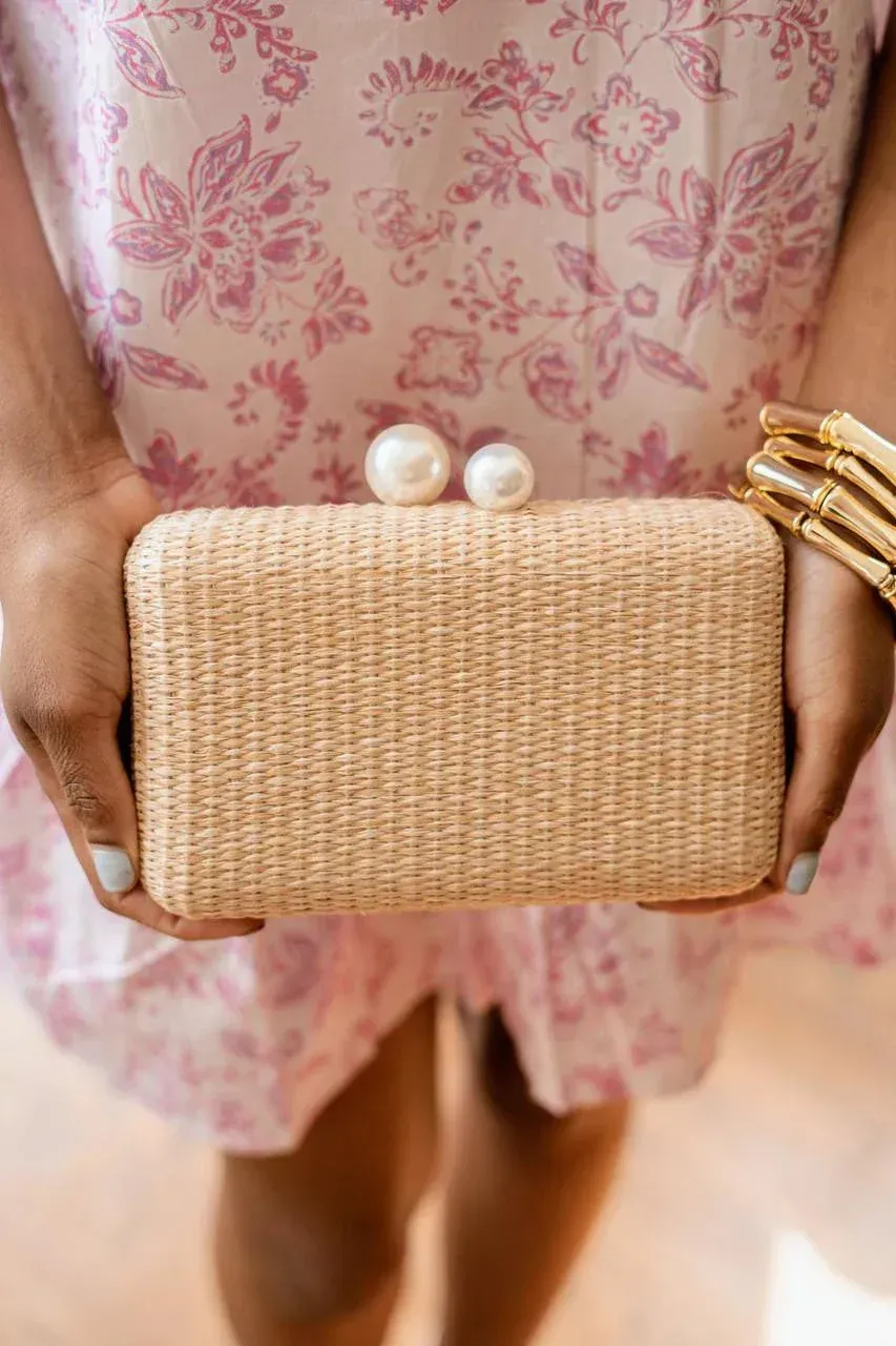 Woman in pink dress holding a beige woven clutch with pearl closure.