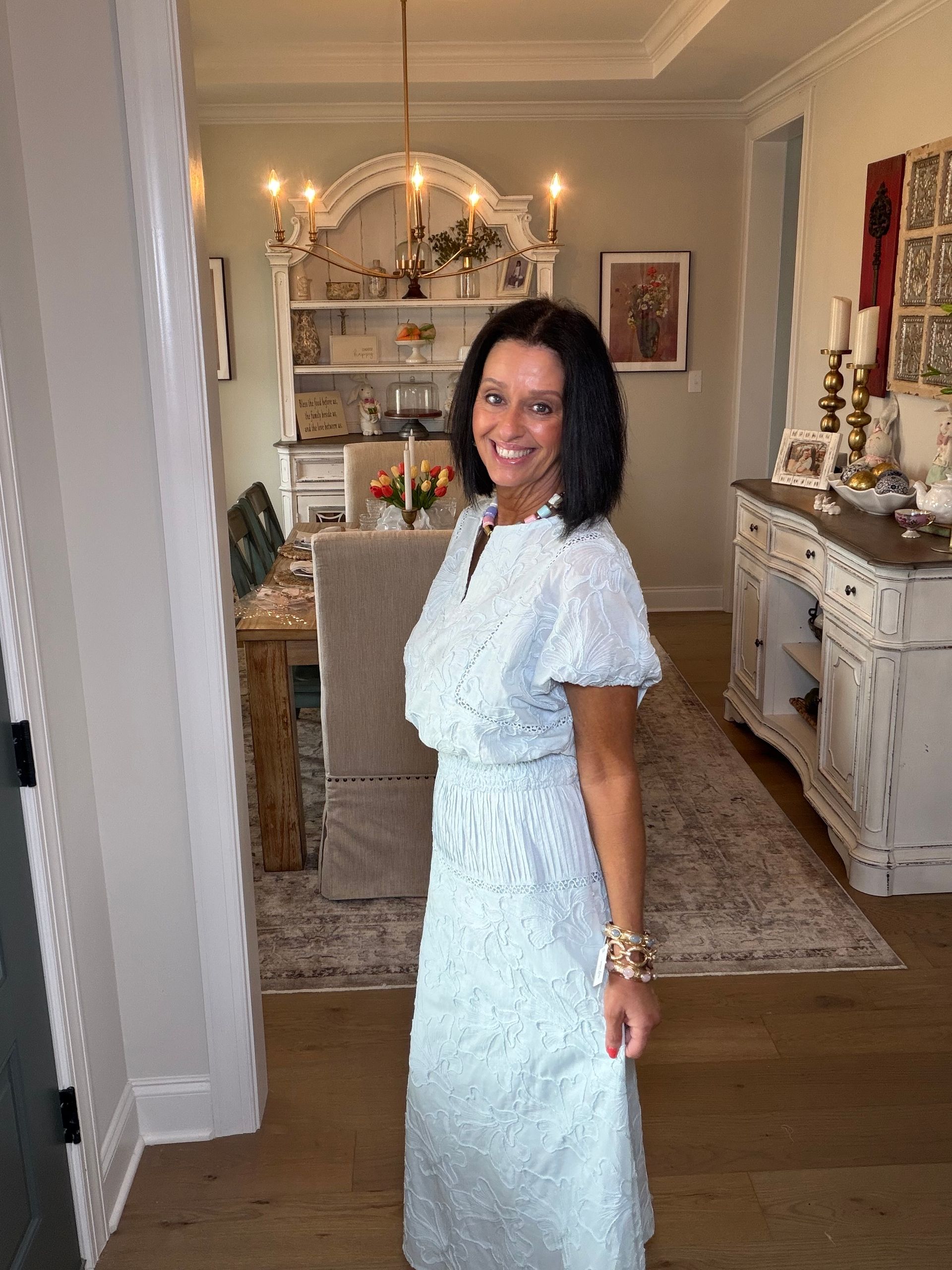 Woman in a beige matching set, short sleeve shirt and wide leg pants, standing in a bedroom.