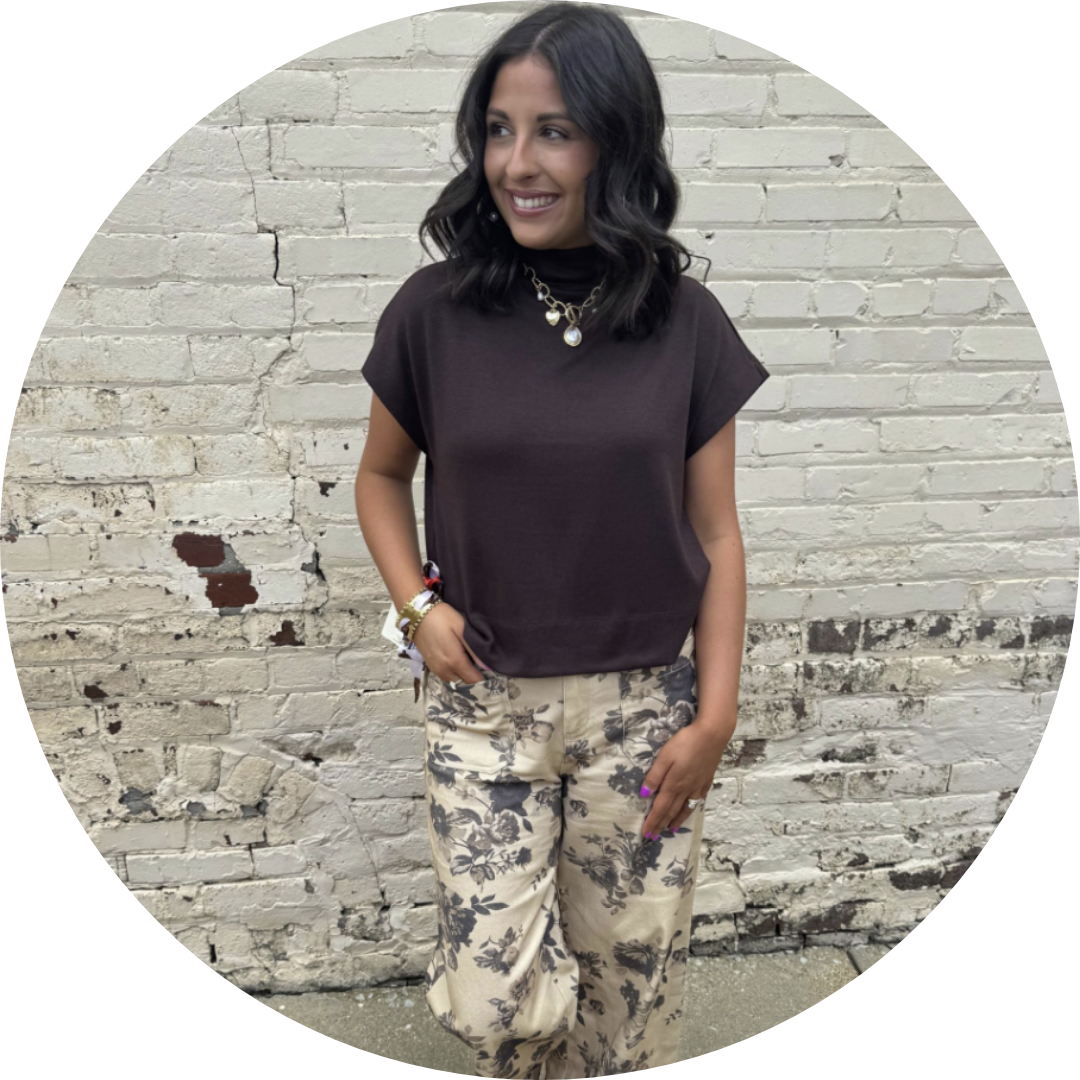 Woman in brown top and floral pants poses against a brick wall.