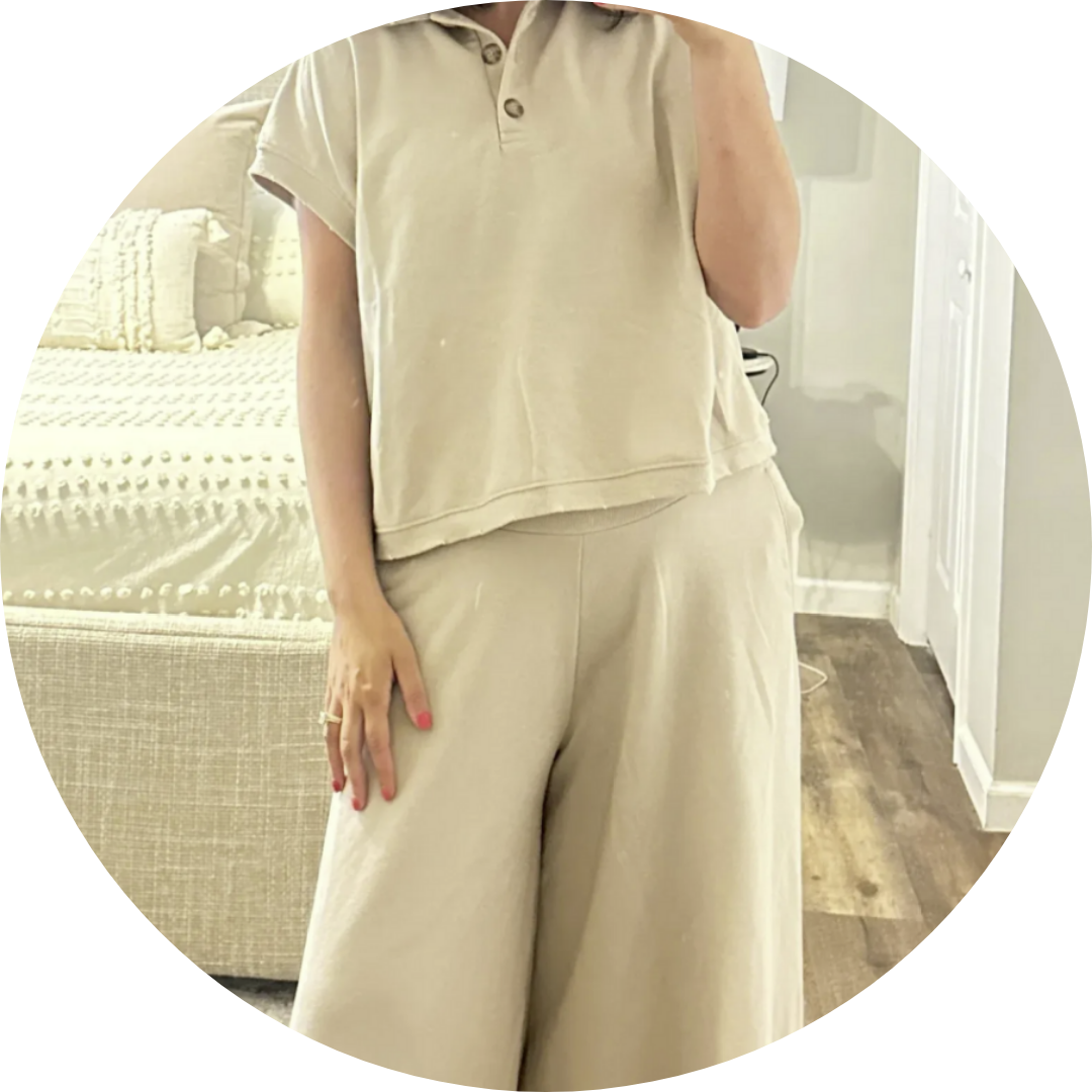 Woman in a beige matching set, short sleeve shirt and wide leg pants, standing in a bedroom.