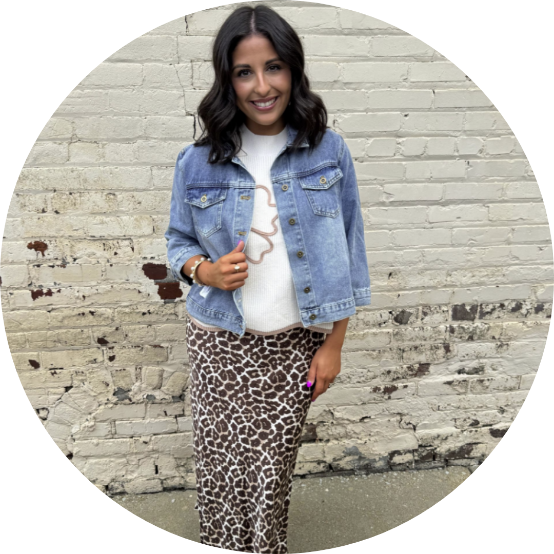 Woman in denim jacket and animal print skirt smiles near brick wall.