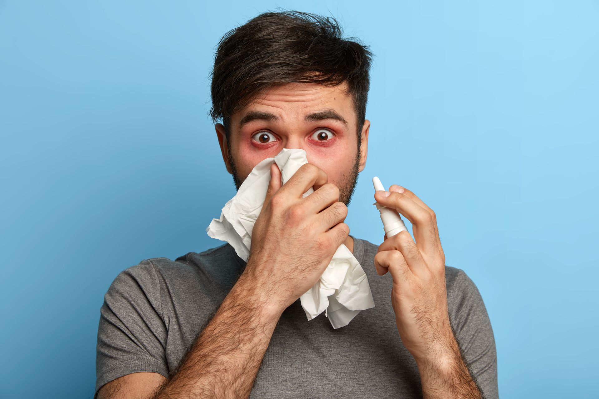 Man holding tissue to nose and eye drops in hand, red eyes, blue background.