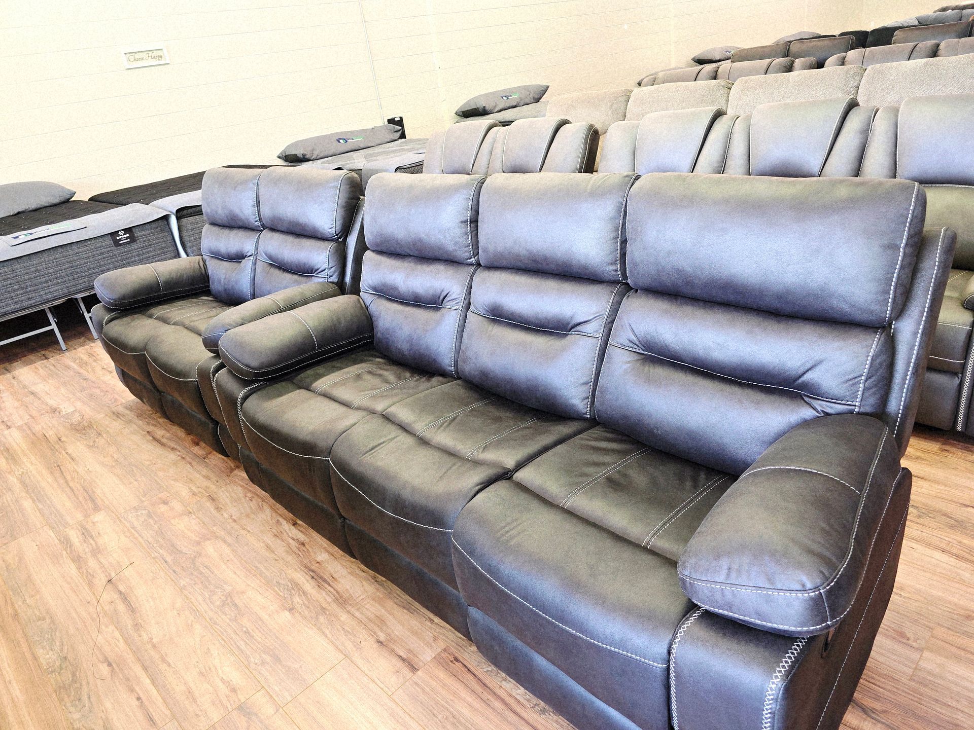 A black leather recliner sofa with white stitching, in a showroom with rows of other furniture.