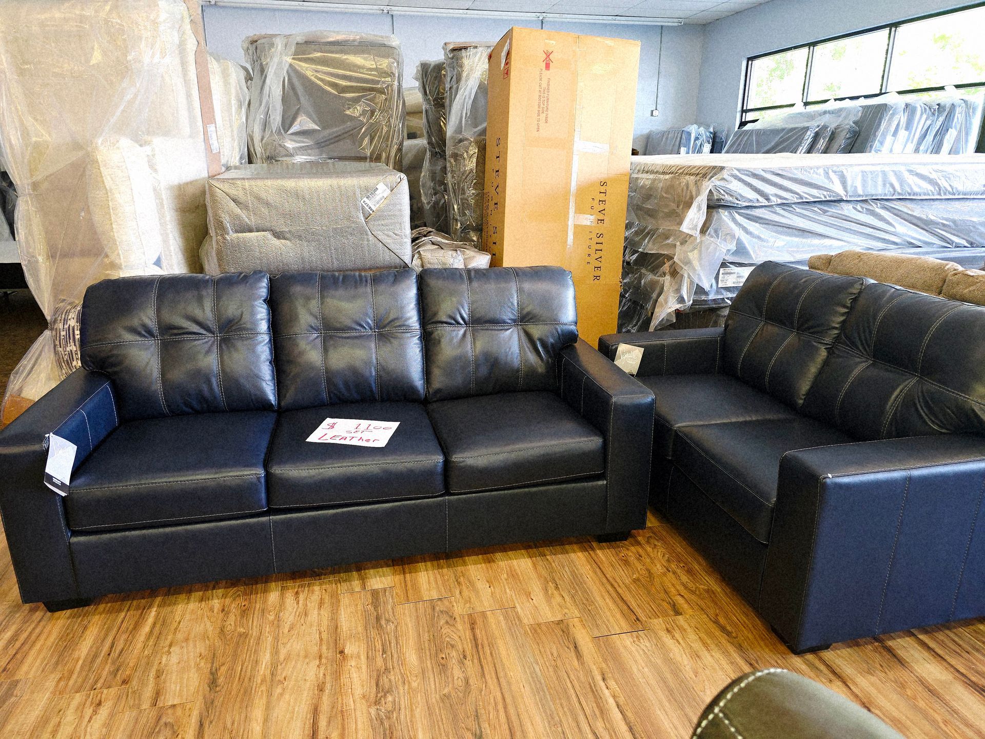 A showroom with a black leather sofa and loveseat set, surrounded by wrapped furniture, on wooden floors.