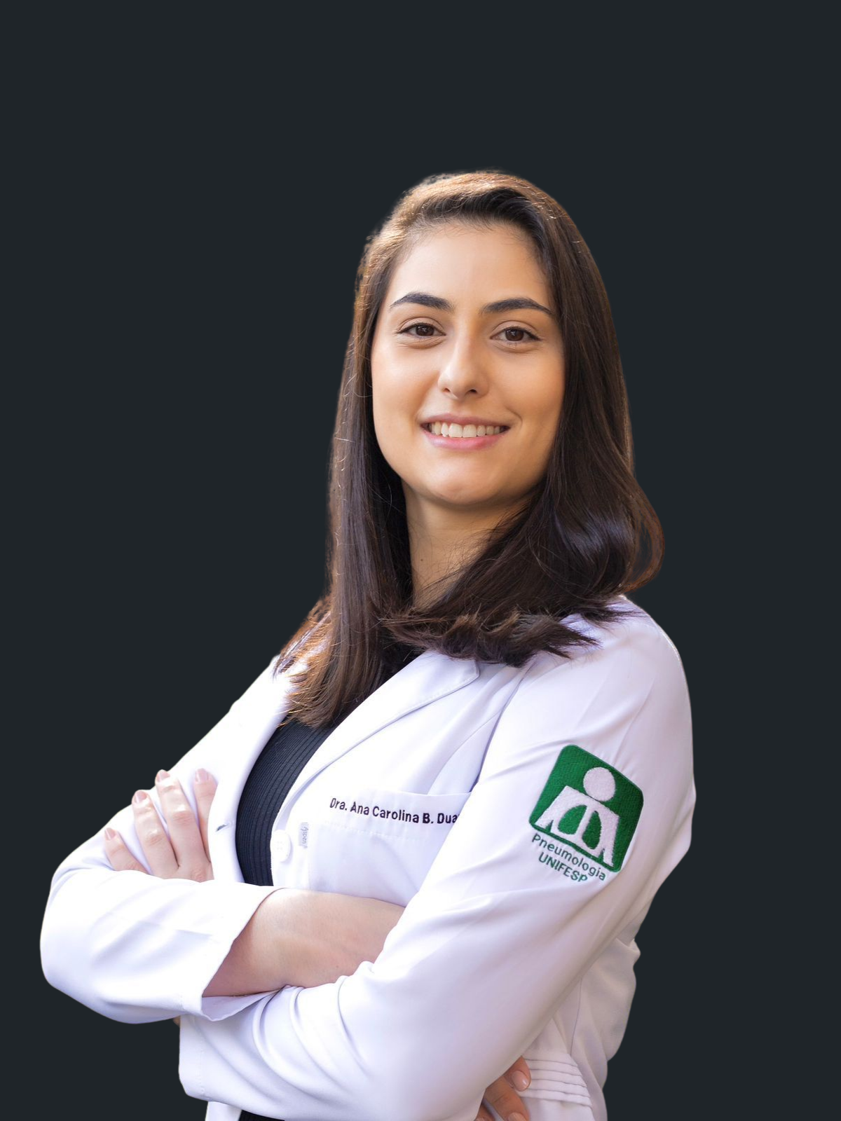 Uma mulher de jaleco está parada em frente a um prédio com os braços cruzados.