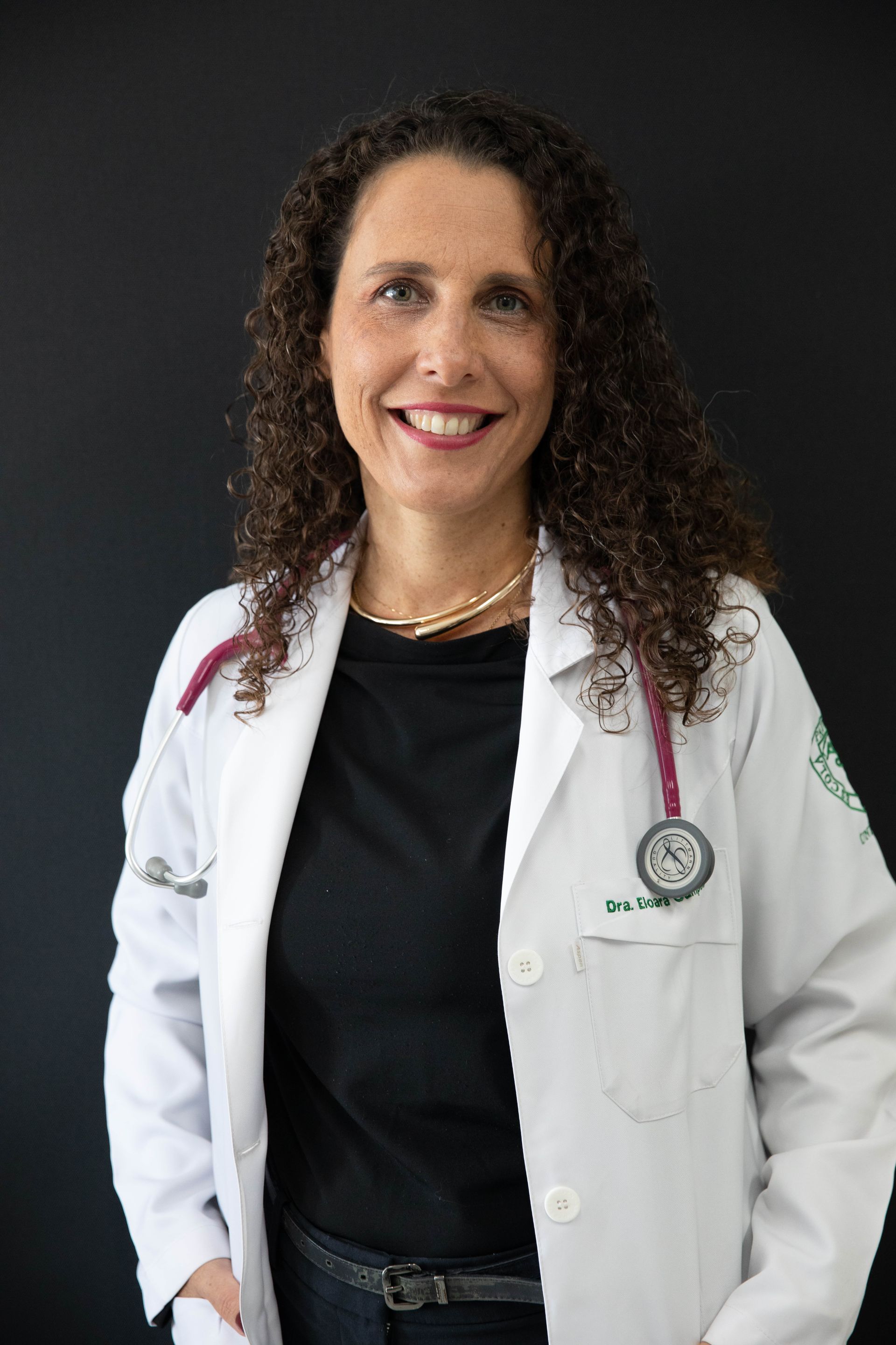 Uma mulher de cabelos cacheados está vestindo um jaleco branco e uma camisa preta.