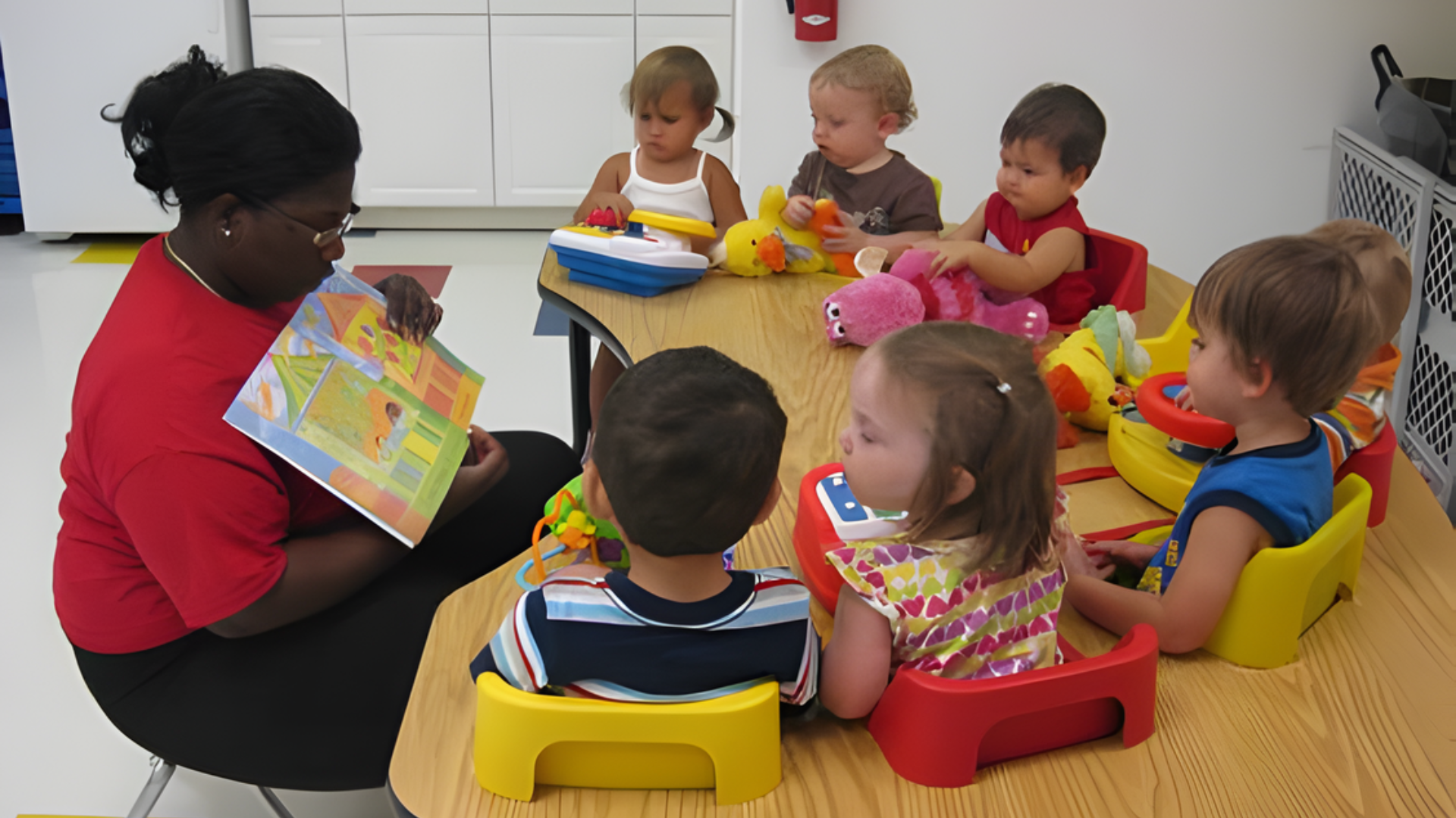 Teacher reading to a group of children seated at a table in a classroom.