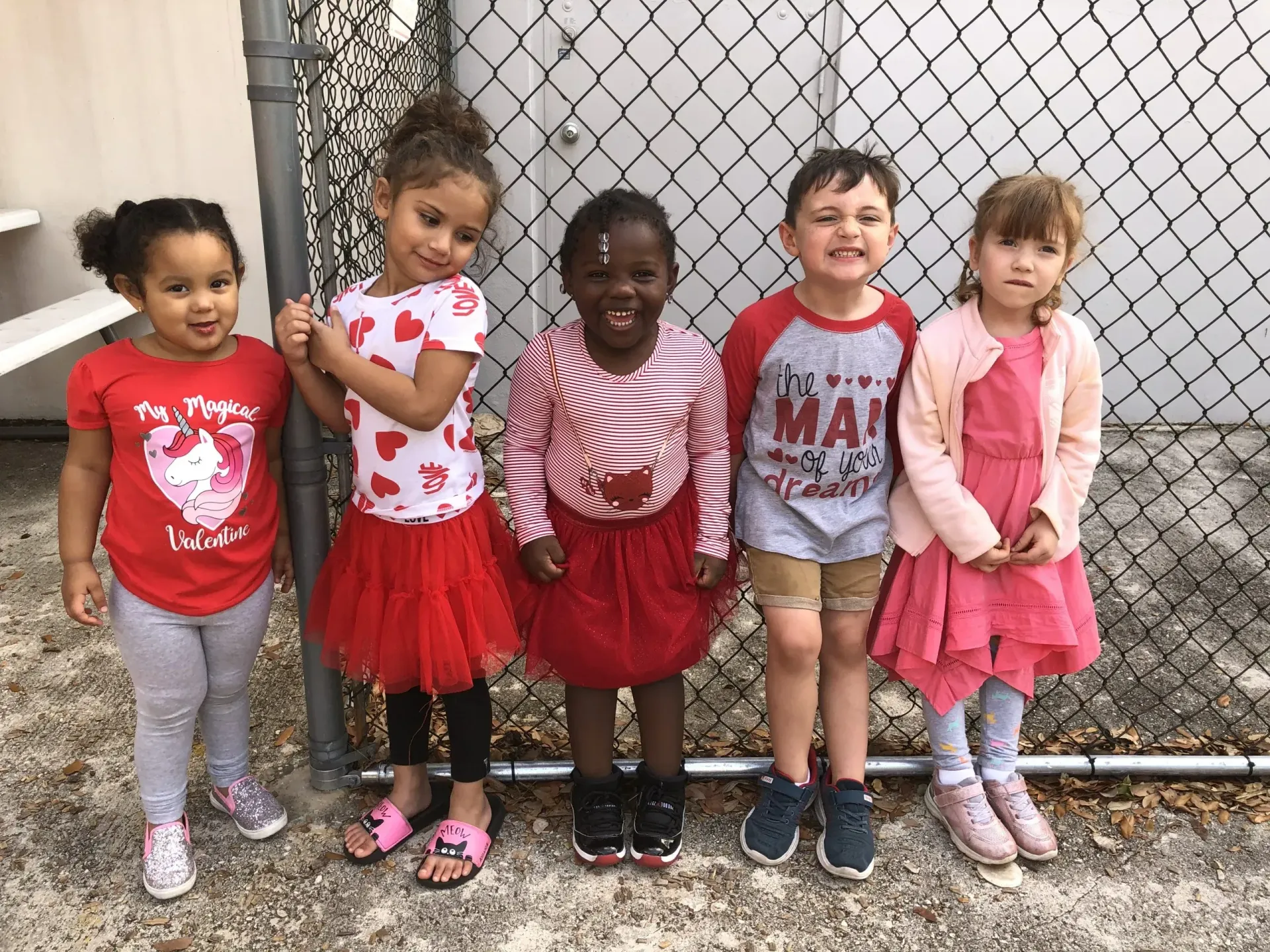Five children in red clothing smiling near a chain-link fence.