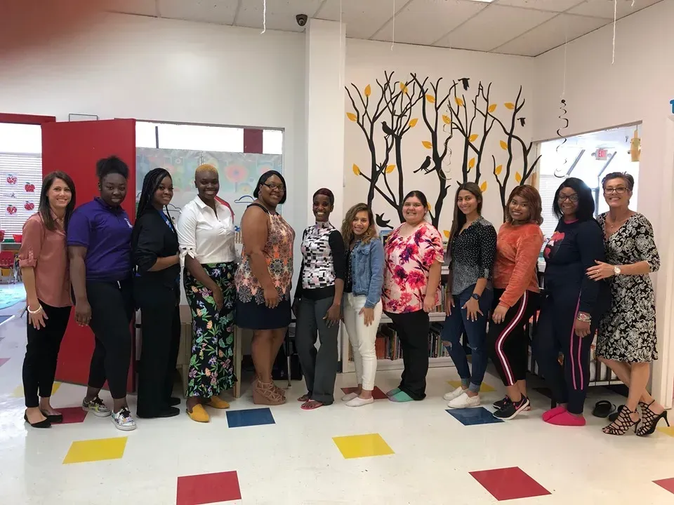 Group of people standing indoors, likely in a classroom, with wall decorations.